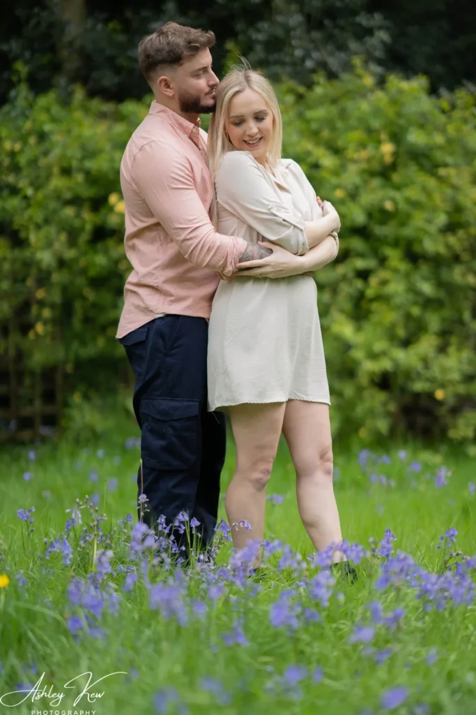 A man and woman stand together in a grassy field with purple flowers. The man, wearing a pink shirt and dark trousers, hugs the woman from behind as she smiles and crosses her arms, with green bushes in the background. Copyright Ashley Kew Photography - All Rights Reserved