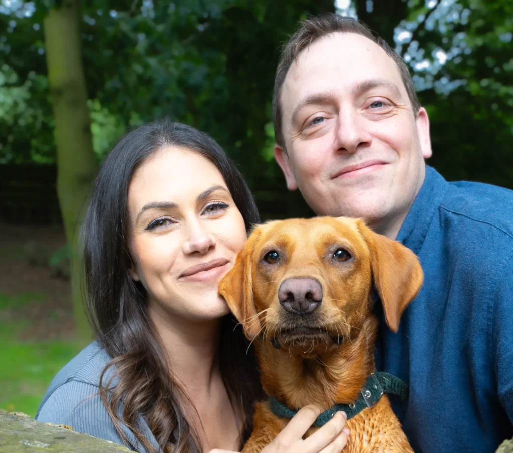 A smiling woman and man pose close together outdoors, holding a brown dog. The trio appear happy, with green trees and foliage blurred in the background. Copyright Ashley Kew Photography - All Rights Reserved