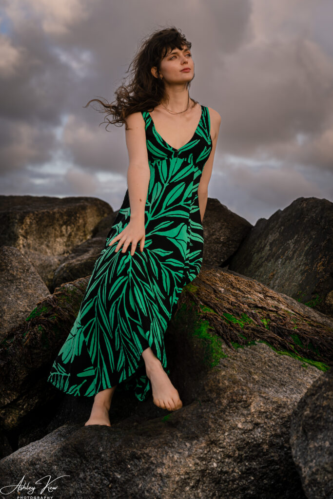 A woman in a green and black patterned dress sits barefoot on large rocks with moss, her hair blowing in the wind under a cloudy sky. Copyright Ashley Kew Photography - All Rights Reserved