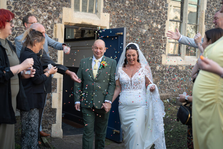 A newlywed couple walks out of a building hand in hand, smiling, as guests on either side throw colourful confetti over them. The bride wears a white dress and veil; the groom is in a green suit. Copyright Ashley Kew Photography - All Rights Reserved