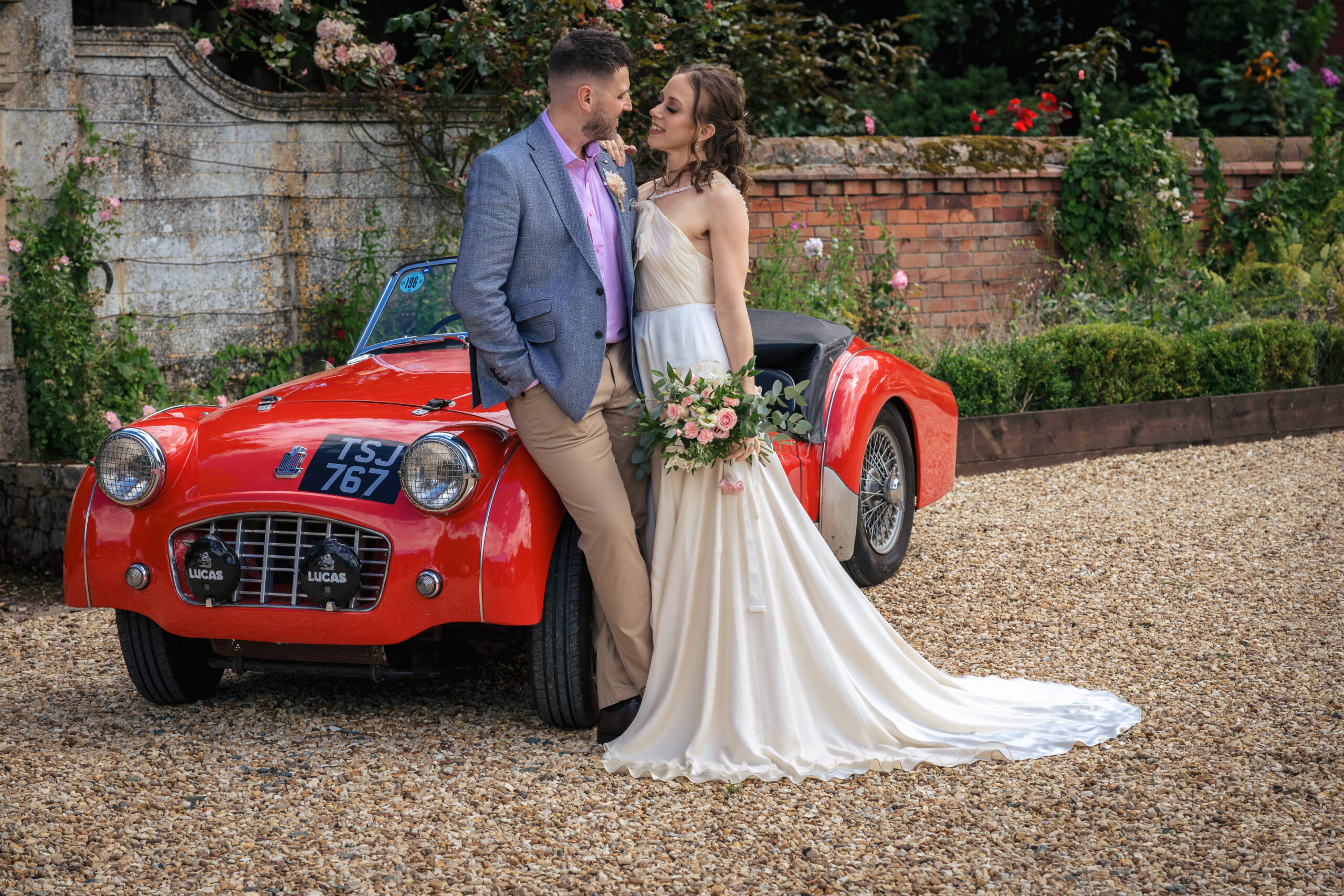 A bride in a long white dress holds a bouquet and stands close to a groom in a light jacket in front of a vintage red convertible car on a gravel drive, with a brick wall and flowers in the background. Copyright Ashley Kew Photography - All Rights Reserved